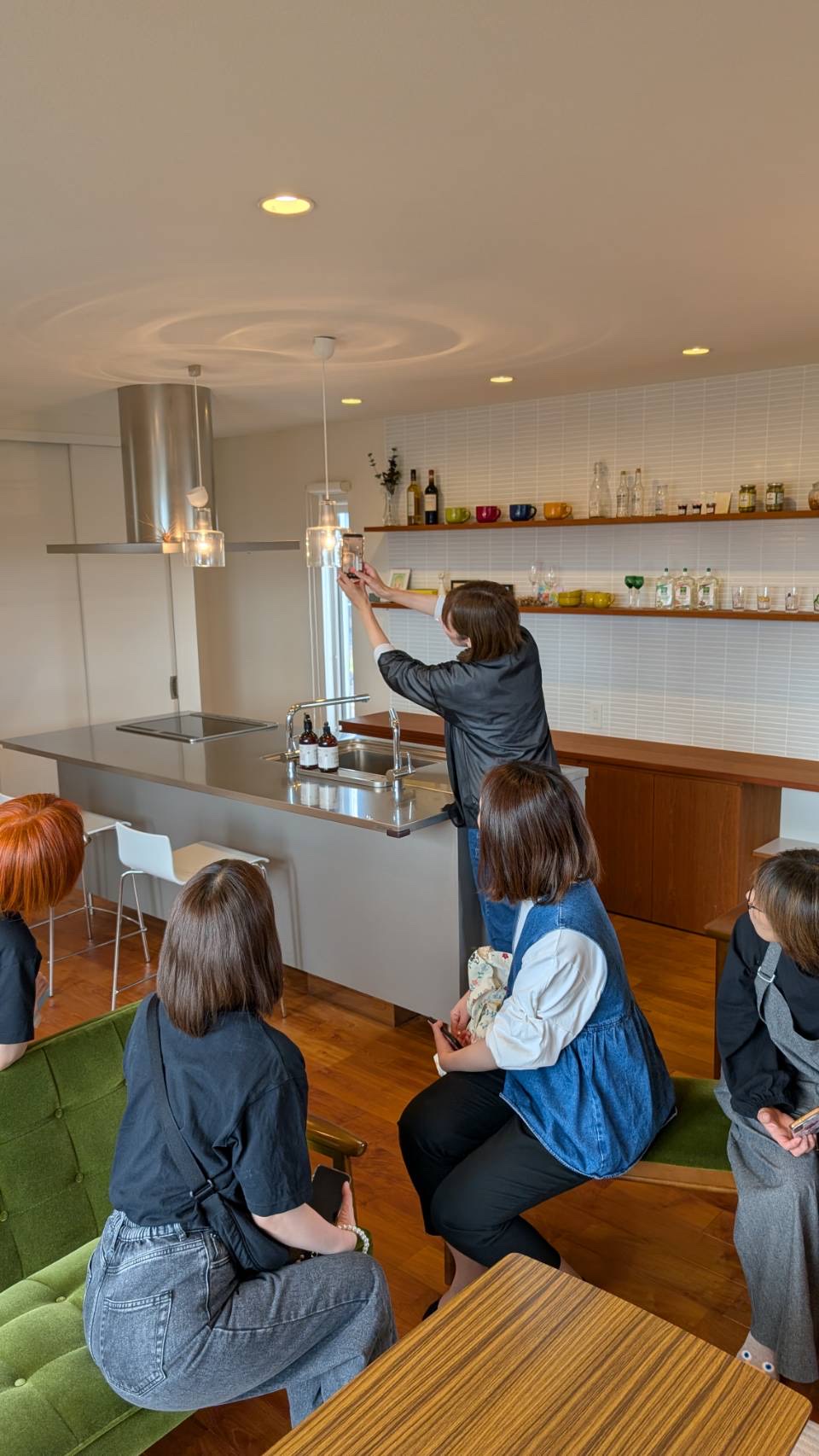 キッチンのある会場で、講師が背伸びしてスマートフォンでの撮影を実演し、参加者が見守る様子